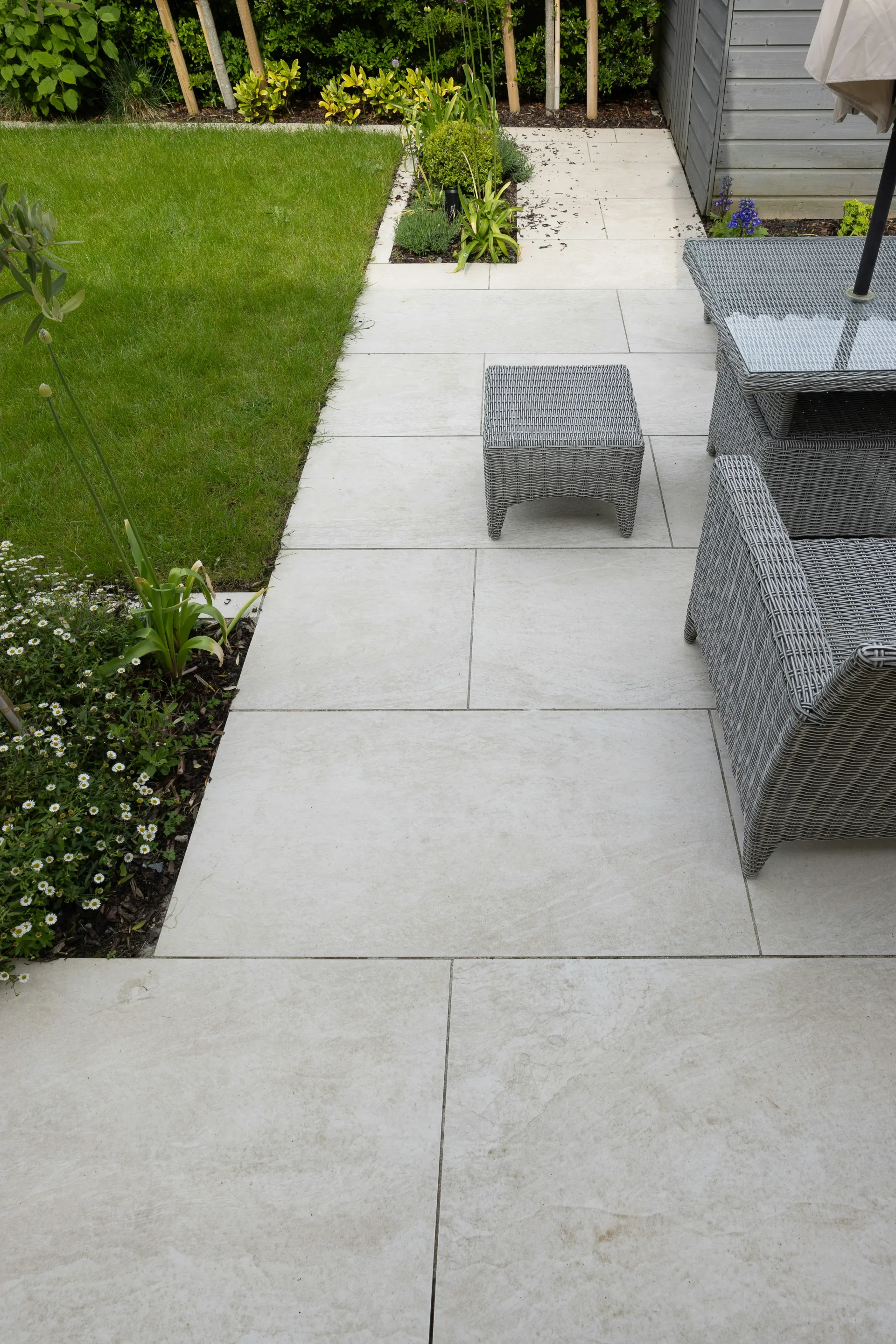 A modern patio laid with large light paving slabs next to a green lawn and garden border with outdoor furniture