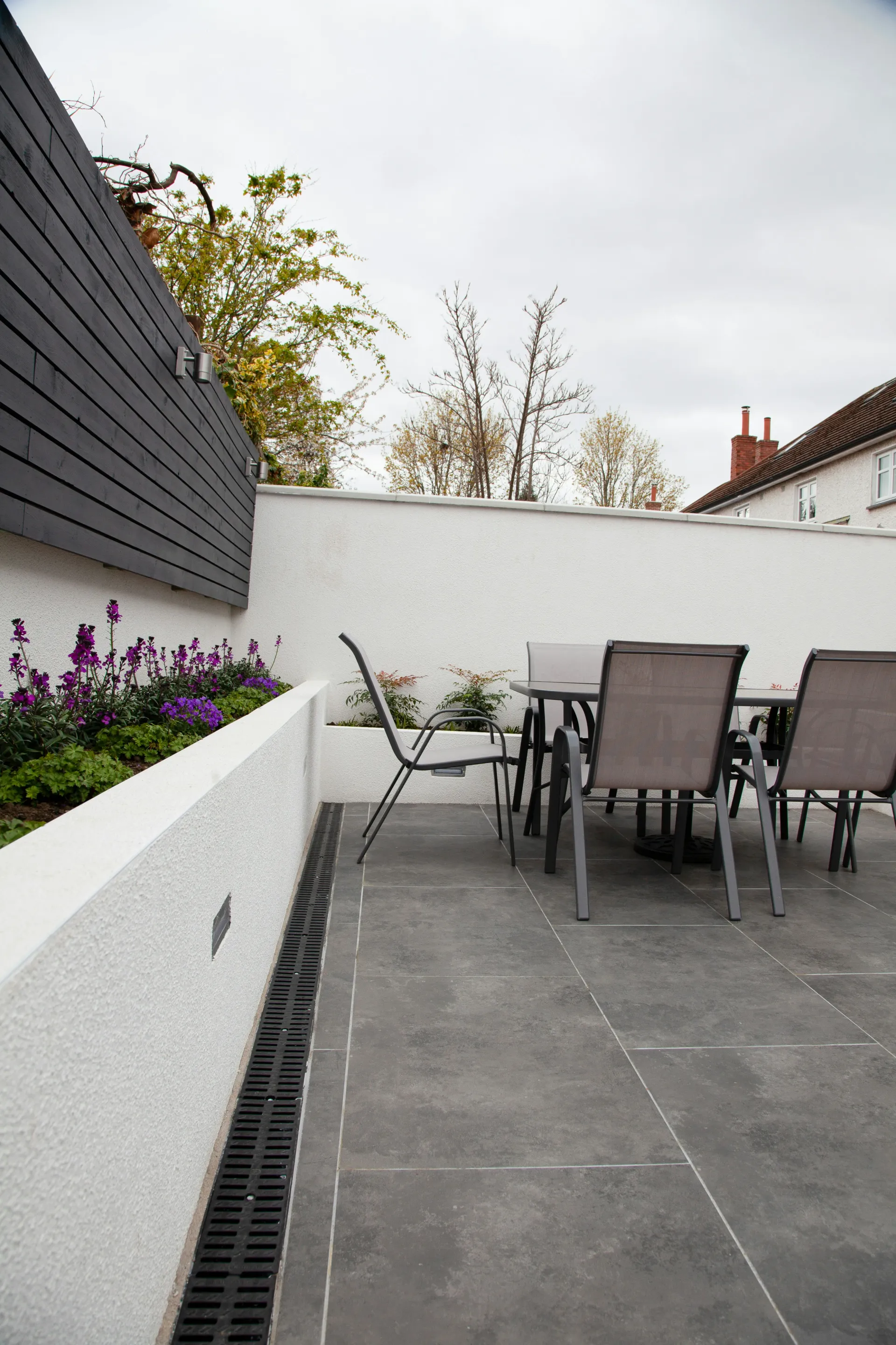 Contemporary grey paved patio with raised planter, drain, and garden furniture.