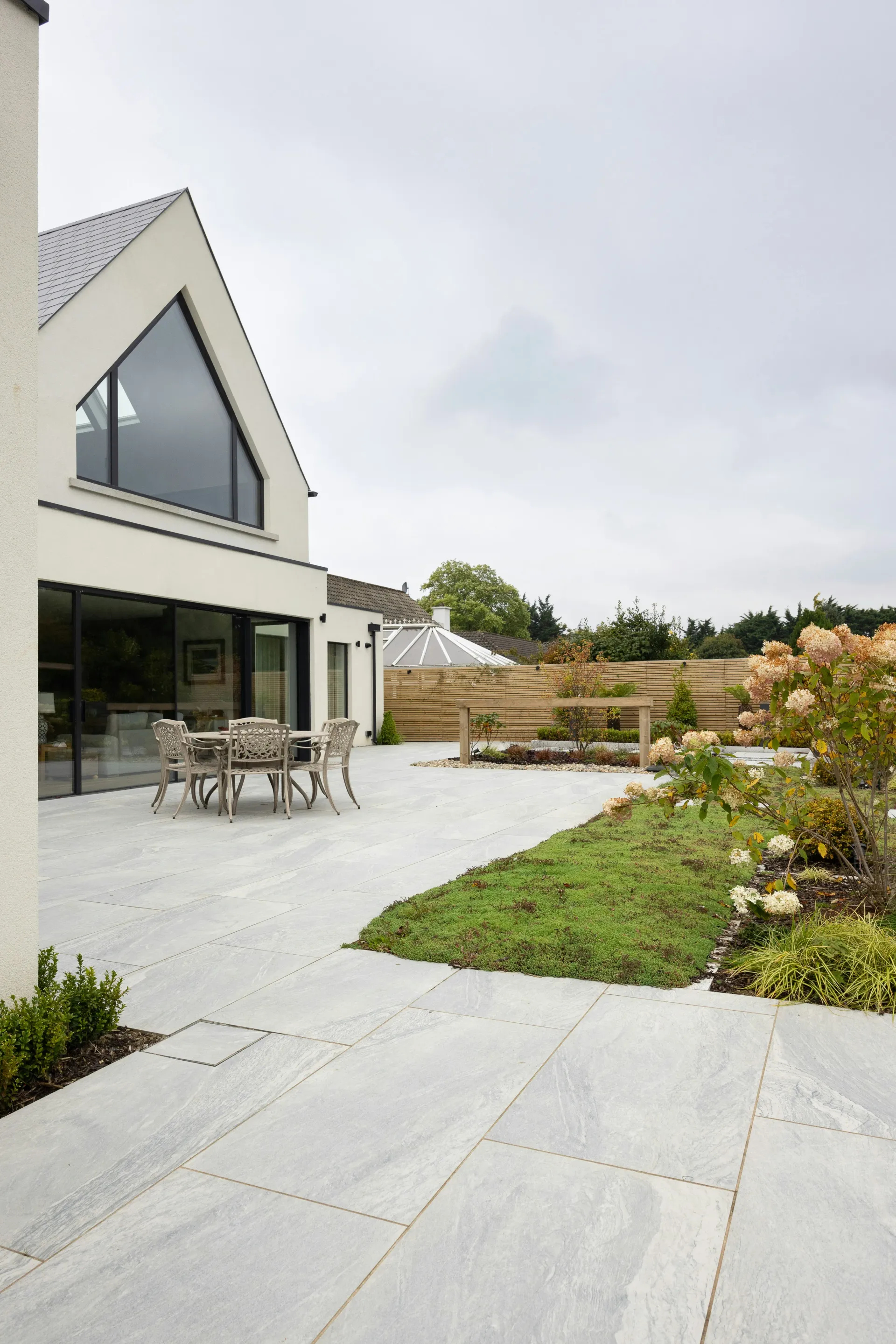 Modern outdoor living space with large grey paved patio, table and chairs, and landscaped garden.